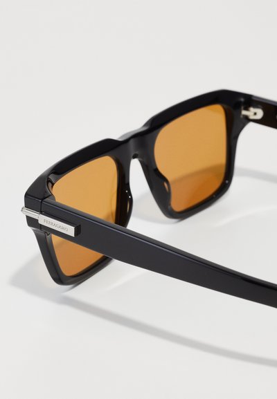 Lunettes de soleil en plastique noir avec des verres ambrés. Elles présentent un design de monture carrée et du matériel argenté avec "Ferragamo" gravé sur la branche.