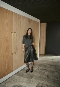 Vrouw met lang, donker haar, gekleed in een grijze trui, zwarte leren rok en zwarte hakken, staand binnen tegen houten kasten.