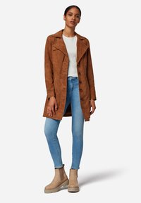 Bruinen suedetrenchcoat met een verschillende kraag, knoopsluiting en zijzakken. Draagt over een witte top en gecombineerd met blauwe skinny jeans en beige laarzen.