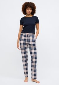 Femme aux cheveux bouclés portant une chemise à manches courtes bleu marine et un pantalon de pyjama à carreaux rose et bleu marine, debout pieds nus sur un fond blanc.