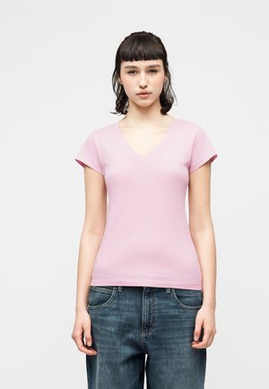 Ung kvinne med kort, mørkt hår iført en lys rosa V-hals T-skjorte og løse blå jeans, stående mot en ensfarget lys bakgrunn.