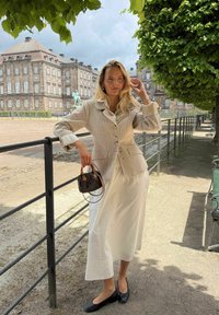 Femme en veste beige et jupe blanche debout près d'une rambarde noire sous des arbres avec un bâtiment historique en arrière-plan par une journée nuageuse.