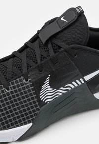 Sort atletik sko med mesh-overdel og gittermønster; har et fremtrædende Nike-logo, justerbar snørelås og tekstureret gummisål.