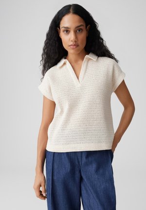 Femme aux longs cheveux bouclés portant un polo en maille crème à manches courtes et un pantalon ample bleu foncé, debout devant un fond uni.