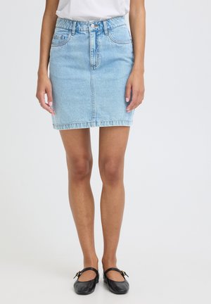 Kobieta w jasnoniebieskiej mini spódnicy dżinsowej, białej bluzce i czarnych płaskich butach Mary Jane stoi na tle jednolitego tła.