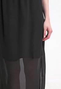 Černé šaty s průsvitným přehozem, které mají rovný střih a boční rozparek. Látka vypadá lehce a má hladkou texturu.