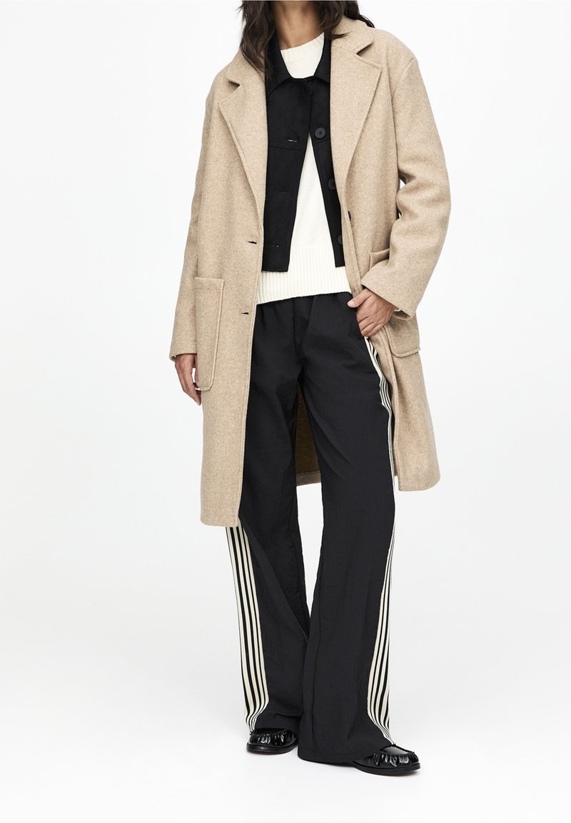 Abrigo de lana beige, chaqueta negra corta, suéter blanco, pantalones negros con rayas blancas, zapatos negros brillantes. Conjunto relajado en capas.