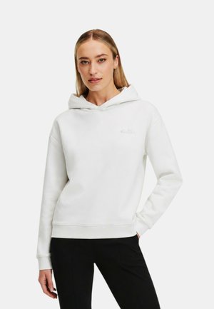 Donna con capelli biondi lisci che indossa una felpa bianca semplice con cappuccio e pantaloni neri, in piedi con una mano nella tasca davanti a uno sfondo bianco.