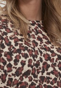 Haut imprimé léopard en beige avec des taches marron foncé et bordeaux. Tissu doux et texturé avec un col rond et des manches courtes.