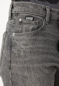 Sive jeans iz denima z rahlo obrabljenim videzom, z žepom na sprednji strani in kovinskimi dodatki. Oznaka G-Star Originals je vidna na pasu.