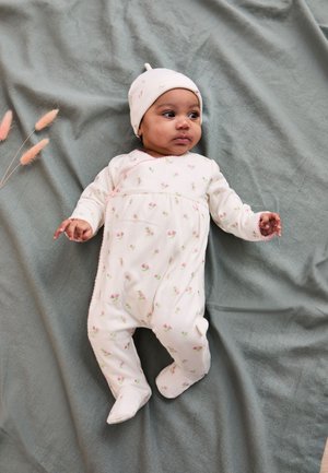 Bábätko leží na sivom deke, oblečené v biele kvetované body a zodpovedajúcu čiapočku, pokojne sa pozerá do strany.