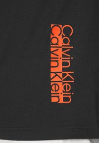 Svart bomullströja med vertikalt orange "Calvin Klein"-logotyp. Korta ärmar, rund halsringning, minimalistisk design, slät textur.
