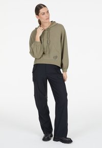 Khakifarbener Kapuzenpullover mit einer Fronttasche und geripptem Saum. Schwarze Cargo-Hose mit mehreren Taschen und einem dezenten Reißverschluss-Detail, kombiniert mit schwarzen Stiefeln.