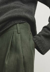 Tummanharmaa neulepaita yhdistettynä vihreisiin laskostettuihin housuihin, jotka korostavat kankaan rakennetta ja rentoa istuvuutta.