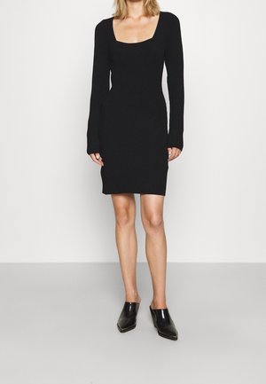 Kobieta ubrana w czarną dopasowaną sukienkę dzianinową z długim rękawem i kwadratowym dekoltem oraz czarne klapki na koturnie z ostrym noskiem, stojąca na jednolitym tle.