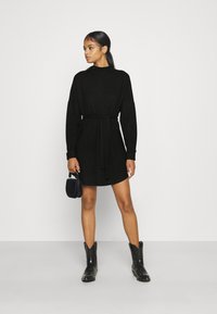 Čierne dlhé šaty s pásom, voľného strihu a vysokým výstrihom, skombinované s čiernymi kovbojskými čižmami a malou kabelkou.