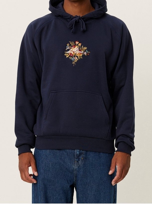 Homme portant un sweat à capuche bleu marine avec broderies florales et texte sur la poitrine, associé à un jean en denim bleu.
