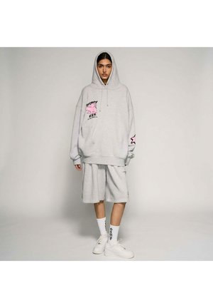 Persona che indossa una felpa con cappuccio oversize grigio chiaro con grafica rosa e pantaloncini lunghi coordinati, calzini bianchi con testo nero e sneakers bianche, in piedi ferma.
