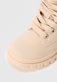 Beige snørestøvler med tykk gummisåle, metallhull og teksturerte vevede snører på glatt lær.