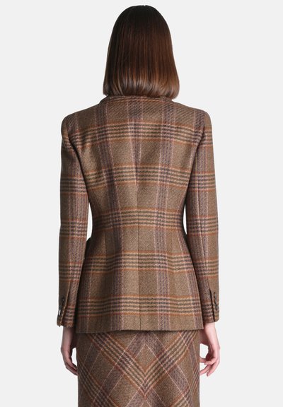 Femme aux cheveux bruns mi-longs portant un blazer marron à carreaux ajusté et une jupe assortie, vue de dos sur un fond uni.