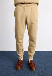 Beige joggebukser laget av mykt stoff med elastisk linning. Har et hvitt Nike-logo og en slank passform. Brukt med brune loafers.