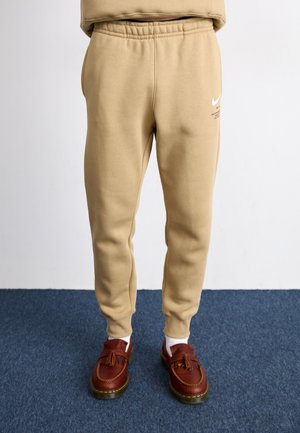 Beige sweatpants made of soft fabric with an elasticated waistband. Features a white Nike logo and slim fit design. Worn with brown loafers.