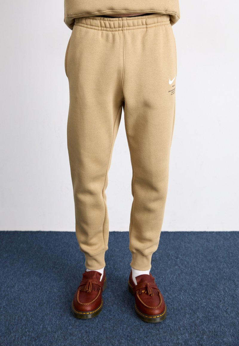 Beige joggebukser laget av mykt stoff med elastisk linning. Har et hvitt Nike-logo og en slank passform. Brukt med brune loafers.