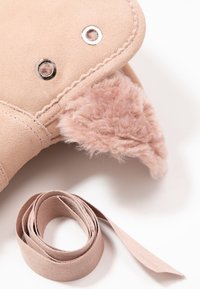 Producto de ante rosa con suaves acentos de piel sintética, detalles de hardware plateado y una correa de tela enrollada al lado. Apariencia texturizada y esponjosa.