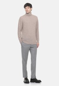 Uomo in piedi che indossa un maglione beige a collo alto, pantaloni grigi e scarpe di pelle nere su uno sfondo bianco uniforme.