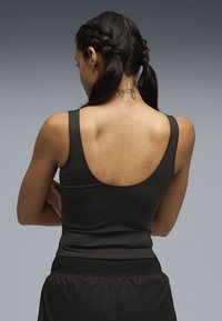 Γυναίκα με πλεξούδες και τατουάζ στο πίσω μέρος του λαιμού, φορώντας μαύρο αμάνικο τοπ και μαύρο παντελόνι, με σταυρωμένα χέρια πίσω από την πλάτη.