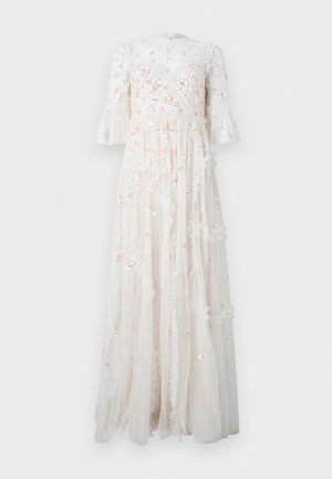 Femme aux cheveux foncés portant une longue robe blanche transparente avec une broderie florale rose et des talons blancs, debout les mains sur les hanches.