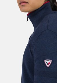 Marineblauer Halb-Reißverschluss-Pullover aus strukturiertem Material, mit einem roten Akzent am Kragen und einem Logopatch auf der linken Schulter.