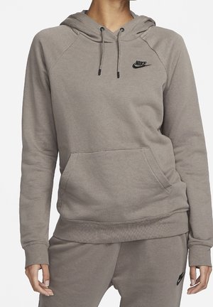 Vrouw die een taupekleurige Nike-hoodie en bijpassende sweatpants met zwarte Nike-logo's draagt, gefotografeerd van bovenlichaam en handen langs het lichaam.