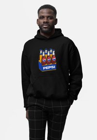 Felpa nera con un grafico di sei bottiglie di soda in una scatola blu etichettata "PEPSI". Il modello indossa pantaloni a scacchi neri con linee bianche.
