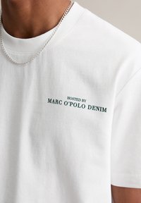 Witte katoenen t-shirt met een geribbelde ronde hals en groene tekst "GEHOST DOOR MARC O'POLO DENIM" aan de linkerkant.