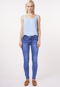 Light blue sleeveless top paired with fitted dark blue jeans. Jeans feature stitching details and five functional pockets, worn with tan slip-on shoes.