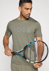 T-shirt de manga curta verde-oliva com logótipo "BOSS", textura lisa; raquete de ténis nas mãos com detalhes em azul e preto e bola de ténis.