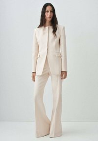 Donna con lunghi capelli scuri che indossa un blazer su misura color crema e pantaloni a gamba larga coordinati, in piedi contro uno sfondo chiaro e uniforme.