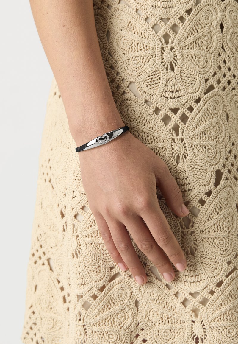 Bracciale elastico nero con accenti argentati, caratterizzato da un design a cuore e piccole pietre incastonate, indossato su una mano contro uno sfondo beige testurizzato.