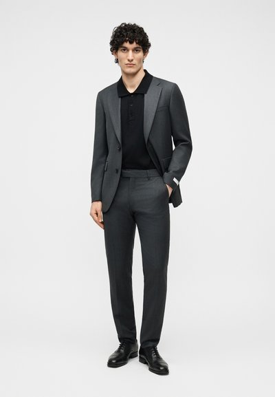 Άνδρας που στέκεται με γκρι κουστούμι, μαύρο πόλο πουκάμισο και μαύρα παπούτσια, με το ένα χέρι στην τσέπη, ουδέτερο φόντο.