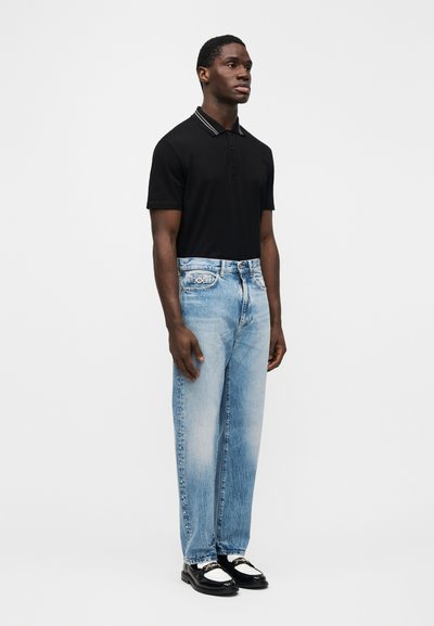 Vyras stovi dėvėdamas juodą trumpomis rankovėmis polo marškinėlį, šviesiai mėlynas išblukusias džinsus su šoniniais kniedėmis ir juodai baltus loaferius, stovėdamas priešais paprastą baltą foną.