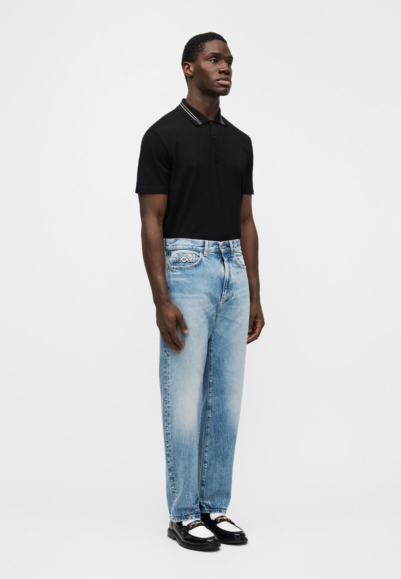 Un homme se tient debout portant un polo noir à manches courtes, un jean bleu clair délavé avec des clous sur le côté, et des mocassins noir et blanc sur un fond blanc uni.