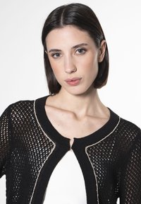 Schwarze Strickjacke mit einer strukturierten, offenen Webart. Auffälliger goldfarbener Besatz entlang des Kragens und der vorderen Öffnung, mit einem runden Ausschnitt.