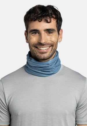 Smiling man with short dark hair wears light grey shirt and blue neck gaiter against plain light background.