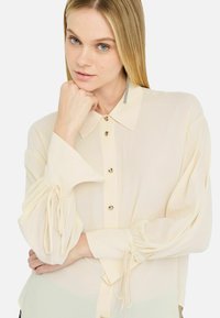 Helle beige Bluse mit durchgehender Knopfleiste, spitzem Kragen und gerafften Ärmeln. Der Stoff wirkt leicht und leicht durchscheinend. Mit Knöpfen akzentuiert.