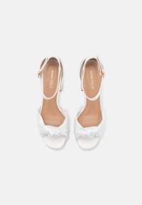 Sandalias de tacón de satén blanco con un diseño anudado en la punta, talón abierto y correa en el tobillo, con una plantilla beige clara y un tacón cuadrado.