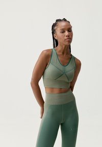 Žalios spalvos sportinė trumpa viršutinė ir aukštai talpūs legginai, pagaminti iš tekstūruoto audinio, su geometriniais raštais ir kontrastingomis apdailos detalėmis.