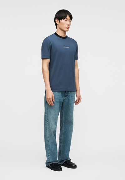 Νεαρός άνδρας στέκεται φορώντας μπλε κοντομάνικη μπλούζα με λογότυπο, ανοιχτόχρωμο μπλε τζιν και μαύρα παπούτσια σε απλό φόντο.