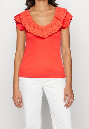 Femme portant un haut rouge vif à manches courtes avec un col en volants festonnés et un pantalon blanc sur un fond uni.