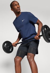 Marineblå Nike T-skjorte med hvit logo, kombinert med svarte shorts med en teksturert panel. Individuell som løfter en vektstang med vektskiver.